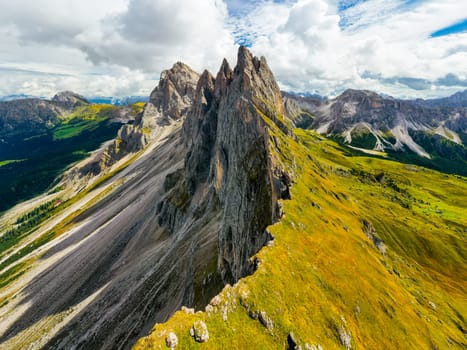 Observation point facing giant Seceda mountain in Italian Alps. Rocky ridgeline in highland under cloudy sky on summer day aerial view