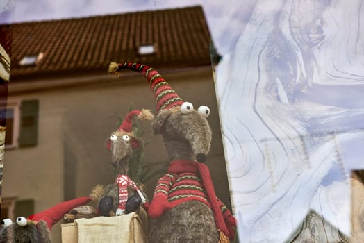 Whimsical New Year's Mouse in Cap and Scarf Stands Behind Store Display. Capture the charm of the holiday season with this delightful image featuring a toy mouse or rat wearing a comical New Year's cap and scarf, playfully standing behind a shop window display. The festive scene adds a touch of whimsy to your creative projects, invoking the spirit of celebration and joy.