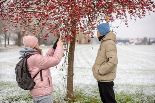 Winter Romance: Girl in Pink Winter Jacket Photographing Boy Against Snow-Covered Red Tree and Field. Embrace the winter magic in this enchanting image, where a girl in a pink winter jacket captures a moment as she photographs her companion against the backdrop of a snow-covered red tree and field. The photograph beautifully conveys the essence of winter romance and the serene beauty of a snowy landscape.