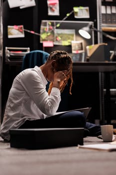 Tired overwored policewoman having headache while analyzing detective report in evening. Exhausted investigator with migraine sitting on floor and reading crime matter file