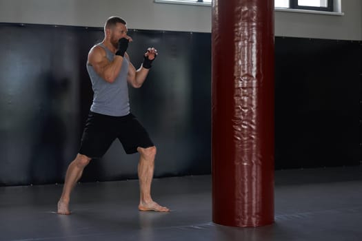 Full body of determined male athlete in sportswear in front of red punching bag. Sportsman with his hands wrapped in boxing tapes in gym during training.