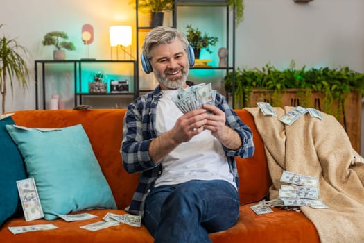 Happy joyful Caucasian senior man in wireless headphones showing wasting, throwing money around, more tips earnings, big profit, win lottery, share, celebrating success on sofa lottery jackpot at home