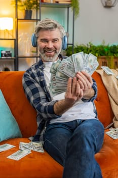 Happy joyful senior man in wireless headphones showing wasting, throwing money around, more tips earnings, big profit, win lottery, share, celebrating success on sofa in living room at home. Vertical