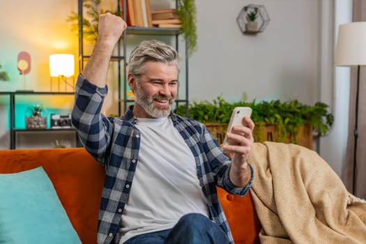 Surprised excited senior man winner holding smartphone reading good news amazed by online bet bid game win. Happy grey-haired Caucasian old guy looking at screen overjoyed by victory success at home.