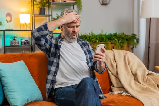 Shocked sad mature man holding smartphone reading negative message feels annoyed sitting on sofa in living room. Caucasian senior elderly old guy having gadget trouble in apartment. App crash concept