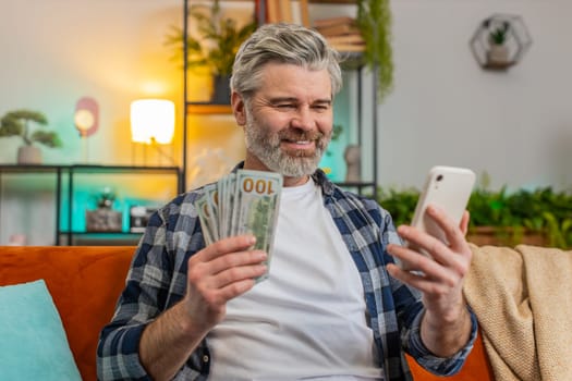 Successful happy rich senior man holding smartphone and counting dollar bill money fan sitting on sofa at home. Happy Caucasian bearded old guy winning online lottery game. Calculating budget concept.