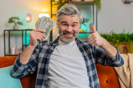 Amazed bearded senior man showing money dollar cash bills clenching at home. Joyful happy middle-aged guy satisfied of income saves pointing money and showing thumbs up. Planning family budget.