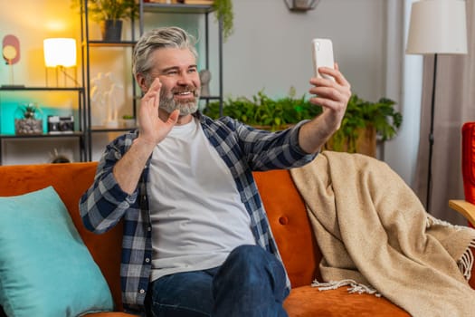 Mature man making video call with wave happy chat and communication on internet online app and social network. Caucasian bearded middle-aged guy using smartphone sitting on sofa in living room at home