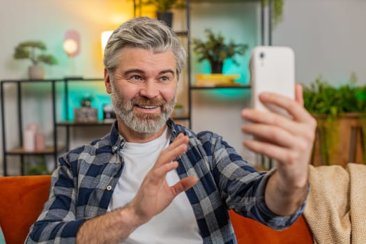 Senior man making video call with wave happy chat communication on internet online app and social network. Caucasian middle-aged grey-haired guy using smartphone sitting on sofa in living room at home