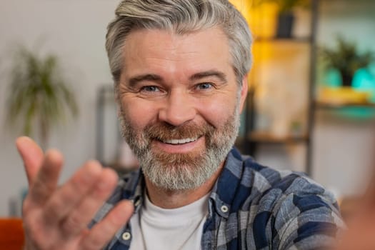 POV portrait of happy senior man in casuals chatting with friend or relative on video online call. Cheerful grey-haired Caucasian middle-aged blogger guy waving goodbye while recording vlog at home