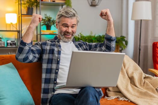 Successful senior man using laptop check e-mail read unbelievable offer, celebrate moment of betting victory feels overjoyed clenched fists. Bearded Caucasian guy working on netbook on sofa at home.