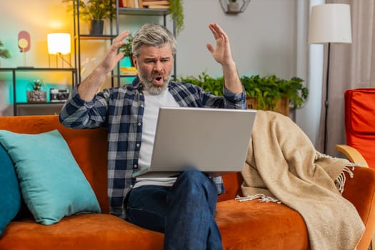 Upset senior man with gray hair sitting on sofa at home uses laptop having anxiety and stress. Shocked Caucasian mature middle-aged guy reacting to loss bad news on netbook in living room in apartment