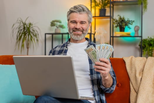 Happy smiling Caucasian senior old man sitting counting money cash and using laptop calculate domestic bills at home. Middle-aged mature guy satisfied of income earnings, saves money, wins the lottery
