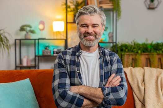 Happy Caucasian senior man sitting on sofa looking at camera and smiling at home. Bearded mature guy having attractive appearance enjoying carefree retired life relaxing in living room in apartment.