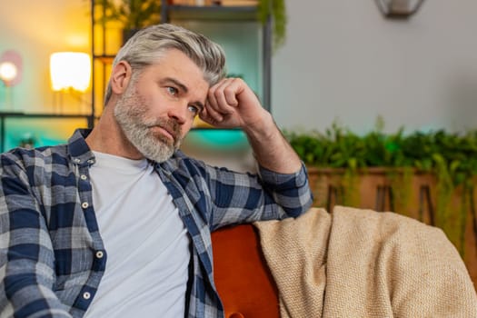 Disappointed man sits at home reflecting on moral distress after quarrel break-up or divorce problem feeling bad annoyed ill sick. Sad middle-aged guy in room contemplates the emotional aftermath