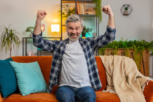 Happy Caucasian senior man watching TV shouting, celebrating success, winning, goal achievement good news, lottery luck, victory sitting on sofa in living room. Mature old guy clenching fists at home.