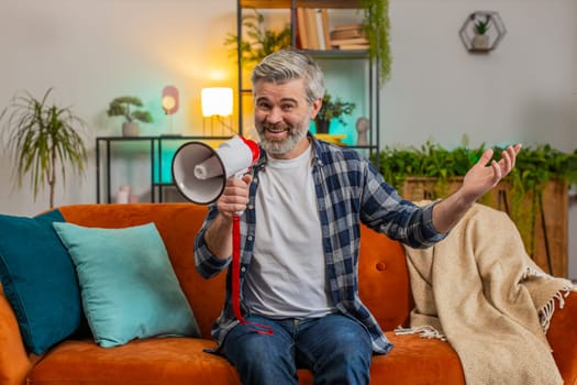 Happy senior man talking into megaphone, proclaiming news, loudly announcing advertisement, warning using loudspeaker to shout speech. Caucasian mature guy announcing big sale sitting on sofa at home