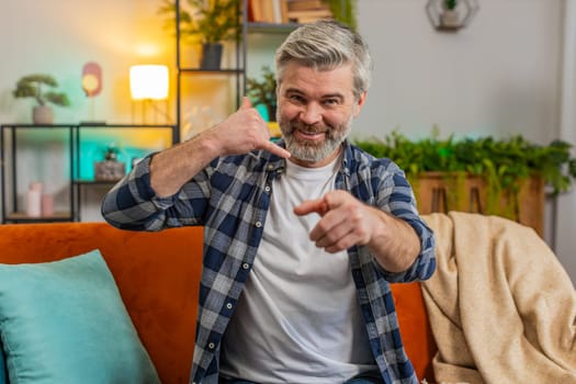 Senior Caucasian bearded old man pointing at camera doing phone gesture saying hey you call me back at home. Mature gray-haired old guy in living room sitting on sofa. Call me, here is contact number