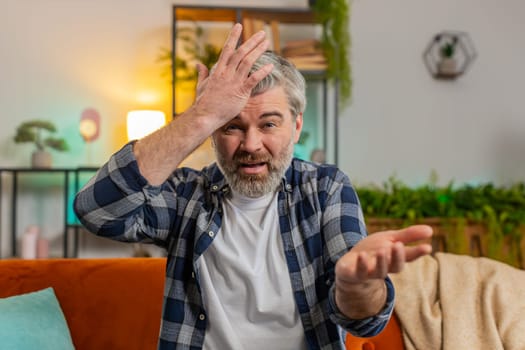 Webcam view. Dissatisfied middle-aged gray haired bearded man arguing quarrel talking on video call sitting on sofa in room at home. Angry mature male guy complains about problems looking in camera