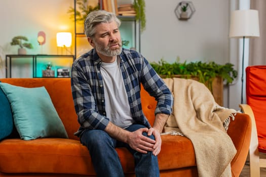 Middle-aged man suffer from painful discomfort severe knee joint ache, leg muscles, could not walk. Sick mature guy massaging knees illness cramps rheumatism at home room on sofa. Health care problem