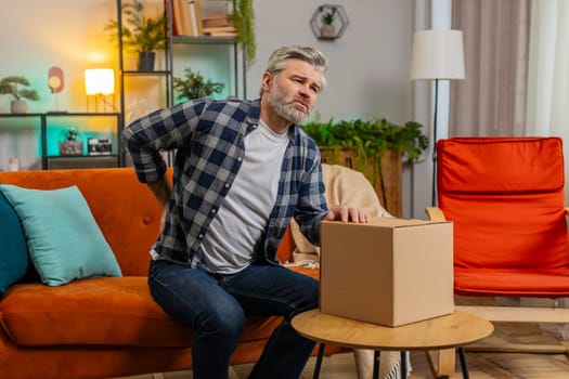 Senior adult man carrying heavy box and experiences severe back pain sitting on sofa in living room at home. Caucasian bearded mature old guy suffering muscle pain of overwork, pinched nerve, spasm