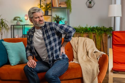 Tired senior bearded man suffering from back pain sitting on couch in living room at home. Sad old male massages painful lower back muscles. Caucasian mature guy exhausted overworked in apartment.