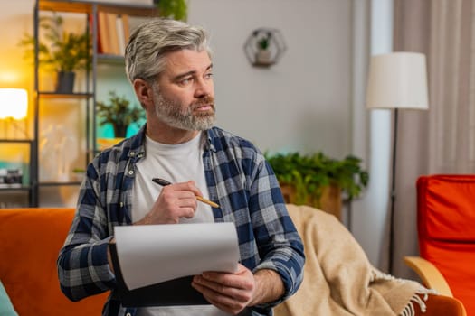 Confident bearded mature man in casual clothes with documents analyzing information reading business papers prepare financial report. Senior Caucasian old guy shocked by sudden victory success on sofa