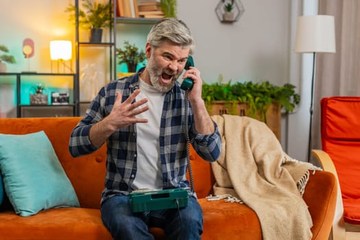 Angry senior man with gray hair talking on vintage retro wired telephone at home apartment. Mad furious Caucasian guy having annoyed boring conversation with hotline helpline service sitting on sofa