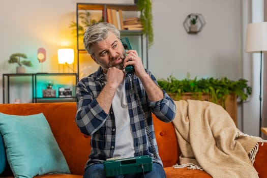 Sad senior man with gray hair talking on vintage retro wired telephone at home apartment. Disinterested Caucasian guy having annoyed boring conversation with hotline helpline service sitting on couch.