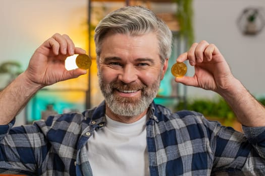 Smiling Caucasian senior freelancer man stock trader holding golden bitcoins BTC near face. Happy excited middle-aged male guy successful developer programmer at home living room. Increasing wealth