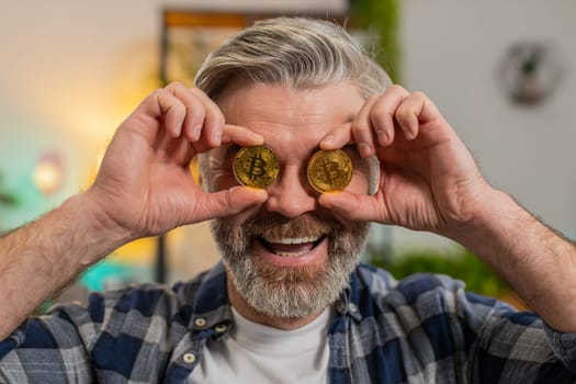 Smiling Caucasian senior freelancer man stock trader holding covering eyes with golden bitcoins BTC. Happy middle-aged male guy successful developer programmer on sofa at home room. Increasing wealth