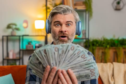 Amazed excited rich joyful Caucasian senior man in wireless headphones showing wasting money cash around more tips earnings, big profit, win lottery jackpot, share, celebrating success on sofa at home