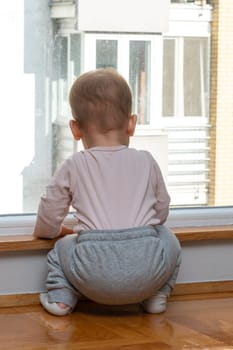 A baby stands near a window in a house, looking at another building and wants to go out for a walk. Concept of feeling alone and wanting to have fun outside