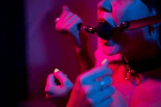 A man gags a woman in a pink blue neon light in the bedroom. BDSM concept