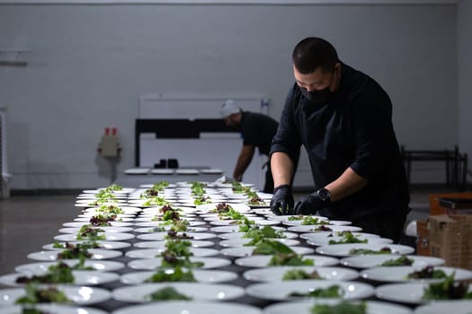 Catering staff wearing gloves and masks prepare plates of fresh salad with efficiency and care for an upcoming event. In the background, tables and chairs are neatly arranged.