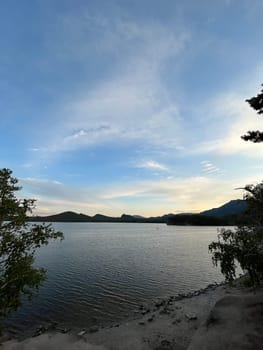 A serene mountain lake at dusk, with the setting sun casting a radiant glow over the tranquil water, creating a picturesque scene with distant hills in the colorful sky.