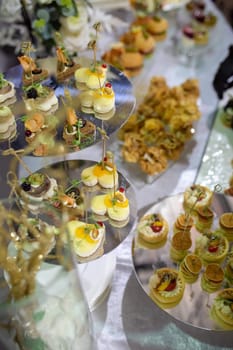 Catering buffet with heart-shaped appetizers, pastries. Smoked salmon blinis, crab cakes, cheese skewers, strawberries, heart-shaped cookies, mini eclairs. Decorated with greenery, flowers.