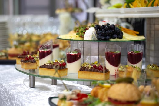 A catering buffet with a variety of delicious snacks and appetizers displayed on glass shelves for a party or corporate event. Includes mini sandwiches, pastries, fruit tarts, cheese, and crackers.