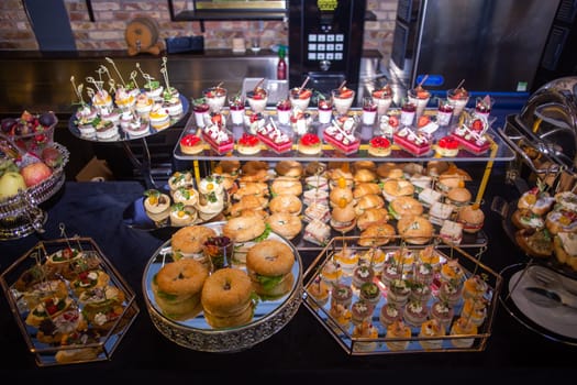 A beautifully presented catering buffet with an array of tempting snacks and desserts. Includes sandwiches, mini burgers, pastries, and fruit, elegantly arranged on tiered trays.