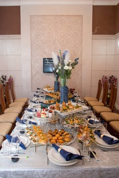 A beautifully arranged table with party food, sandwiches, pastries, fruits, and vegetables. Stylish chairs and a striking floral centerpiece create a sophisticated ambiance for a special occasion.