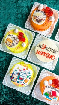 An assortment of six small square cakes with unique icing designs and decorations in plastic containers on a green surface, featuring cartoon characters, strawberries, and other decorative elements.