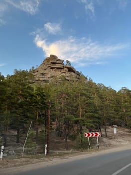 Towering rock formation emerges from the forest, commanding attention with its unique shape. Surrounded by greenery, rugged surface bears witness to time, sky adorned with wispy clouds.