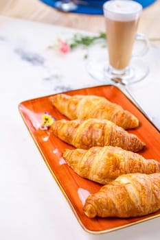 Delicious breakfast of golden croissants, coffee, and flowers on a white marble table. Croissants arranged on orange plate with coffee cup and flowers on right. Blurred background.