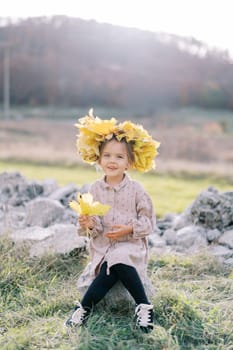 Little smiling girl in a wreath of autumn leaves sits on a stone on the lawn with yellow leaves in her hand. High quality photo