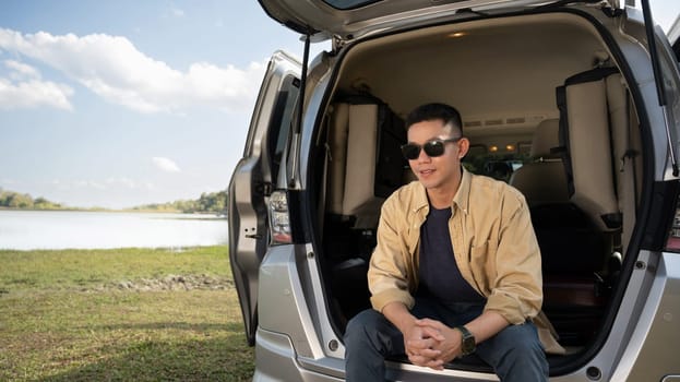 Relaxed young man in sunglasses sitting in the open trunk of minivan near lake with mountains and clear sky.