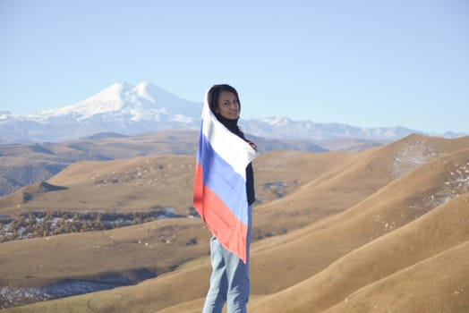 A young brunette woman stands against the backdrop of the snow-capped Mount Elbrus, looking at the camera, a Russian flag covers her shoulders. Tricolor against the backdrop of snow-capped Mount Elbrus.
