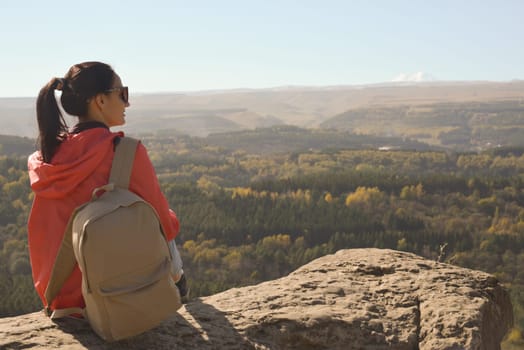 A girl tourist with a backpack sits on the top of a mountain and enjoys the beautiful views. A walking woman in an orange jacket rests on a rock, looking at the beautiful sunny landscape of the Caucasus Mountains