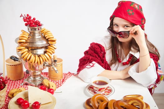 A fashionable modern girl in stylized folk clothes at table with a samovar, bagels and tea for the Orthodox holiday of Maslenitsa and Easter. Funny photo shoot for a young woman