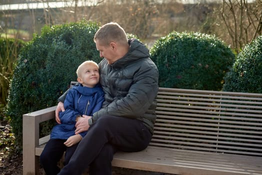 Autumnal Family Affection: Father, 40 Years Old, and Son - Beautiful 8-Year-Old Boy, Seated in the Park. Embrace the warmth of familial love in the autumnal air with this heartwarming image. A father, 40 years old, and his son - a beautiful 8-year-old boy, seated in the park. The essence of autumn adds a touch of seasonal charm to this delightful moment of family affection.