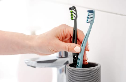 Girl'S Hand Places Two Toothbrushes In Cup For Hygiene In Bright Bathroom
